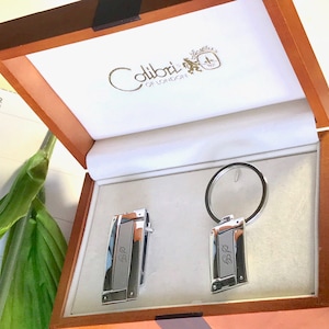 May include: A wooden box with a white interior, containing a silver key chain and cufflinks. The keychain and cufflinks are engraved with the initials "B.P.". The box is labeled "Colibri of London".