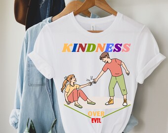 May include: White t-shirt featuring a colourful "KINDNESS OVER EVIL" graphic. The graphic depicts two cartoon figures, one extending a hand to the other.