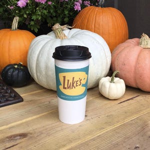 May include: A white takeaway coffee cup with a black lid and a green and yellow logo that says "Luke's" with a stylised coffee cup underneath. The logo also says "Stars Hollow, Connecticut". The cup is sitting on a wooden table with several pumpkins in the background.