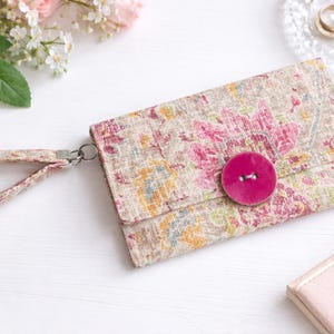 May include: Floral print clutch with a wrist strap, featuring a large pink button closure. The fabric has a beige background with pink, yellow, and green floral patterns. A silver-tone clasp connects the strap to the clutch.