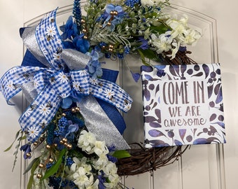 Op de afbeelding: Een blauwe en witte krans met een bord waarop staat "Come in we are awesome". De krans is versierd met blauwe en witte bloemen, een blauw-wit geruit lint en zilveren glitter.