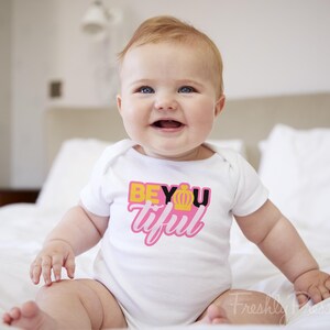 May include: A white short-sleeved baby bodysuit featuring a graphic print on the chest. The text reads BEYOUtiful in yellow, black, and pink lettering, with a small yellow crown icon replacing the letter O in the word BEYOU.