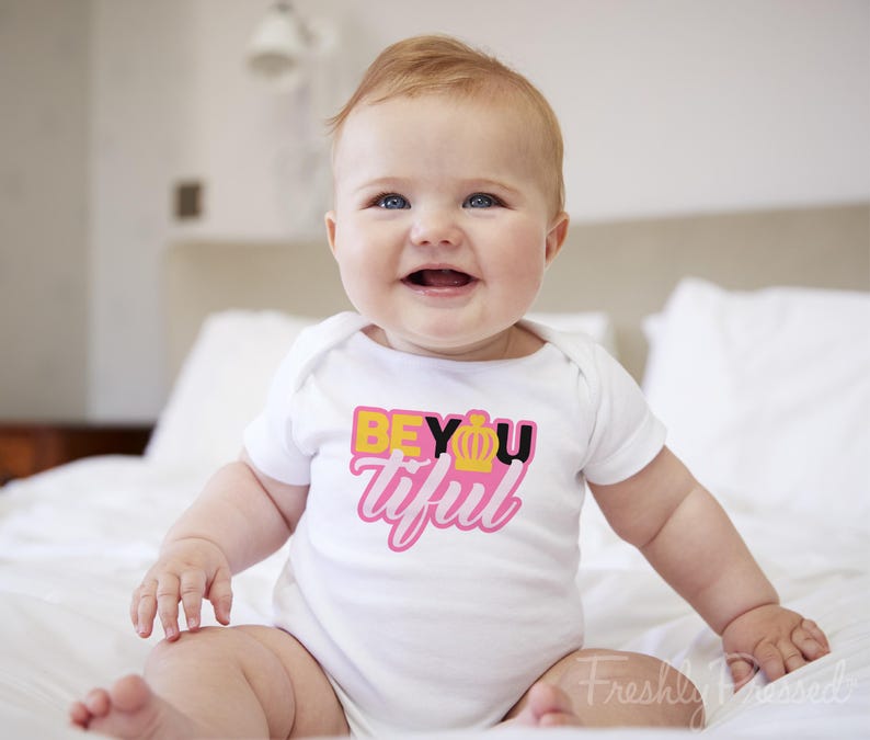 May include: A white short-sleeved baby bodysuit featuring a graphic print on the chest. The text reads BEYOUtiful in yellow, black, and pink lettering, with a small yellow crown icon replacing the letter O in the word BEYOU.