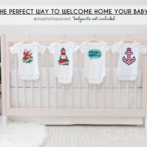 May include: Four white baby onesies hanging on a white crib with nautical themed designs. The onesies feature a red bird with a banner that says "Welcome Home", a red and white lighthouse with the text "Sail Away", a speech bubble with the text "Feelin' Nauti", and a red and white anchor.