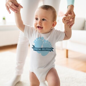 K&ouml;nnte beinhalten: Ein wei&szlig;er Baby-Strampler mit einem blauen Baby-Fu&szlig;abdruck-Design und dem Text "SPECIAL DELIVERY" auf einem blauen Band.