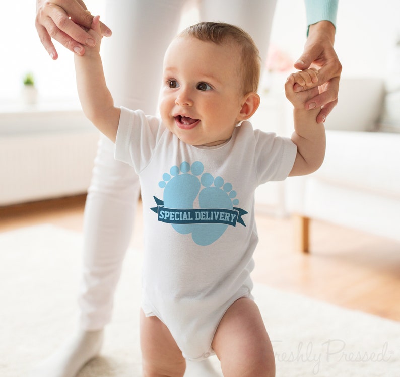 K&ouml;nnte beinhalten: Ein wei&szlig;er Baby-Strampler mit einem blauen Baby-Fu&szlig;abdruck-Design und dem Text "SPECIAL DELIVERY" auf einem blauen Band.
