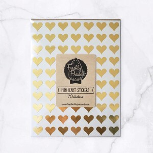 May include: A package of 70 mini heart stickers in various shades of gold. The stickers are arranged on a white background. A brown label with the text "Freshly Pressed" and "Mini Heart Stickers" is in the center.