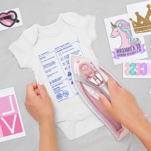 May include: A white baby bodysuit being ironed with a pink and grey iron. The bodysuit features a printed instruction sheet. Surrounding the bodysuit are various iron-on transfer designs, including heart-shaped glasses, a unicorn, a gold crown, and text.