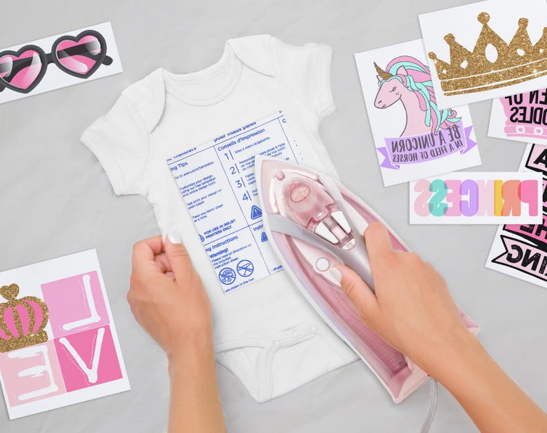 May include: A white baby bodysuit being ironed with a pink and grey iron. The bodysuit features a printed instruction sheet. Surrounding the bodysuit are various iron-on transfer designs, including heart-shaped glasses, a unicorn, a gold crown, and text.