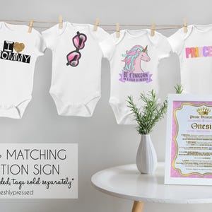 May include: Six white baby bodysuits hanging on a clothesline, each featuring unique graphic prints including a crown, heart, sunglasses, unicorn, the word PRINCESS, and a fox. A framed instruction sign sits on a white table next to a small plant.