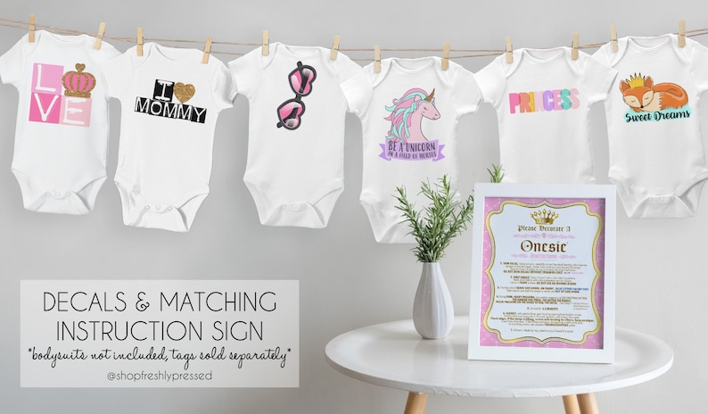 May include: Six white baby bodysuits hanging on a clothesline, each featuring unique graphic prints including a crown, heart, sunglasses, unicorn, the word PRINCESS, and a fox. A framed instruction sign sits on a white table next to a small plant.