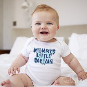 May include: A white baby onesie with a blue and white graphic that says "Mommy's Little Captain" with a nautical anchor and a seashell.