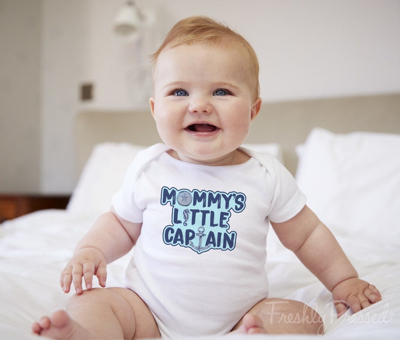 May include: A white baby onesie with a blue and white graphic that says "Mommy's Little Captain" with a nautical anchor and a seashell.