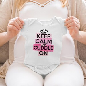 May include: A white short-sleeved baby bodysuit featuring a black crown graphic above the text "KEEP CALM AND CUDDLE ON" printed in black letters on pink rectangular backgrounds. The garment has a round neckline and snap closures at the bottom.