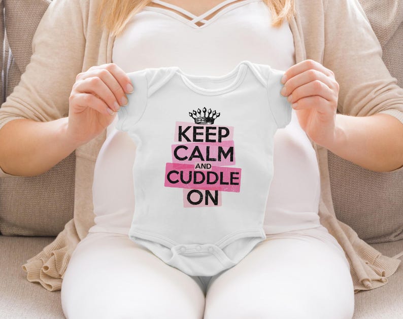 May include: A white short-sleeved baby bodysuit featuring a black crown graphic above the text "KEEP CALM AND CUDDLE ON" printed in black letters on pink rectangular backgrounds. The garment has a round neckline and snap closures at the bottom.