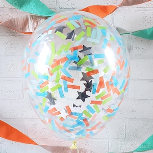 May include: A clear balloon filled with colorful confetti and star-shaped accents. The confetti includes orange, blue, green, and black rectangular pieces. The balloon is tied with a yellow ribbon, and streamers in orange and teal are in the background.