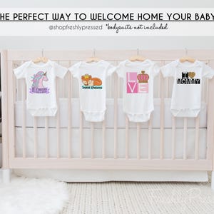 May include: Four white baby bodysuits hanging on wooden hangers. The first features a unicorn and text, the second a fox with a crown and text, the third the word LOVE with a crown, and the fourth the text I heart MOMMY with a glitter heart.