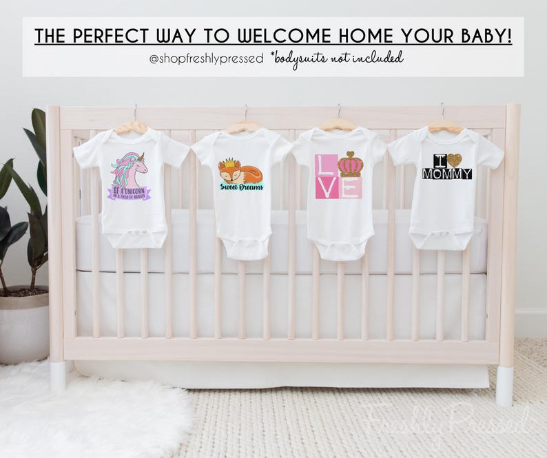 May include: Four white baby bodysuits hanging on wooden hangers. The first features a unicorn and text, the second a fox with a crown and text, the third the word LOVE with a crown, and the fourth the text I heart MOMMY with a glitter heart.
