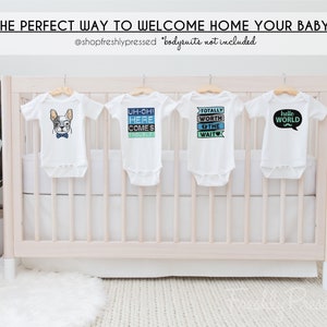 May include: Four white baby onesies hanging on a white crib. The onesies have different designs, including a cartoon dog, the words "Uh-oh! Here comes trouble!" and "Totally worth the wait", and a speech bubble with the words "Hello World" and a mustache.