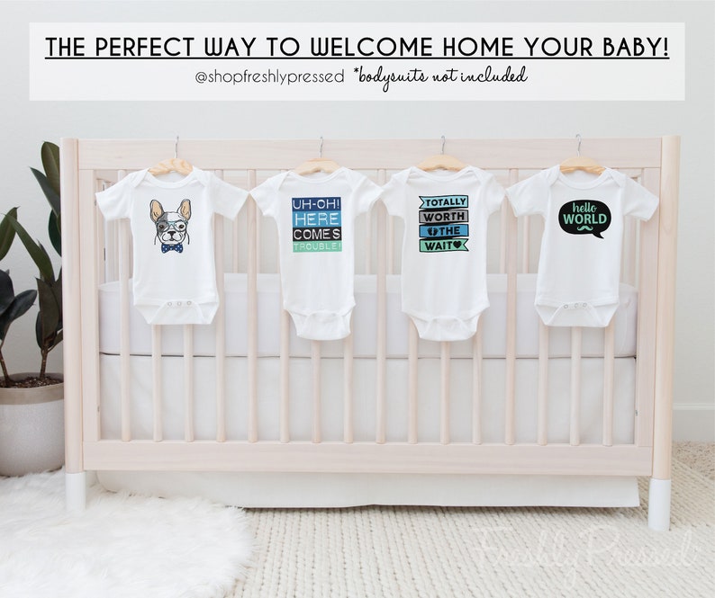 May include: Four white baby onesies hanging on a white crib. The onesies have different designs, including a cartoon dog, the words "Uh-oh! Here comes trouble!" and "Totally worth the wait", and a speech bubble with the words "Hello World" and a mustache.