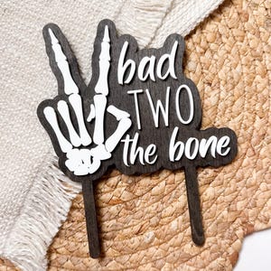 May include: A wooden cake topper with a dark brown background and white text that reads "bad TWO the bone." The design includes a white skeleton hand making a peace sign. Two brown sticks extend from the bottom for insertion into a cake.
