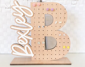 May include: Wooden earring holder shaped like the letter B with the word "Bexley" written in white on the side. The holder has multiple holes to display earrings.