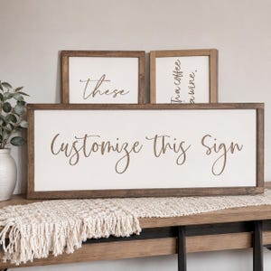 May include: Three rectangular wooden signs with white backgrounds and dark brown frames. The large horizontal sign features the text "Customize This Sign" in brown script. Two smaller vertical signs are behind it with partial text visible.