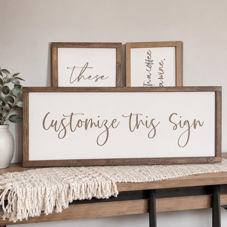 May include: Three rectangular wooden signs with white backgrounds and dark brown frames. The large horizontal sign features the text "Customize This Sign" in brown script. Two smaller vertical signs are behind it with partial text visible.