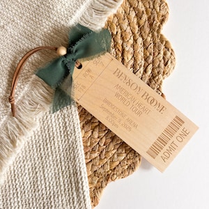 May include: A rectangular wooden ticket ornament engraved with Benson Boone American Heart World Tour details, including Bridgestone Arena, September 9, 2025, 8:00pm, seat 7, row A, and a barcode. It features a dark green chiffon ribbon bow and a leather cord.