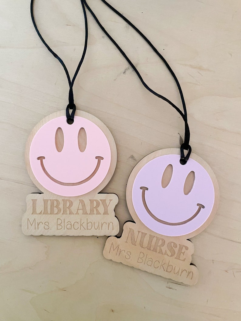 Pu&ograve; includere: Due targhette in legno con facce sorridenti e lacci neri. La targhetta superiore dice "LIBRARY Mrs. Blackburn" e la targhetta inferiore dice "NURSE Mrs. Blackburn".