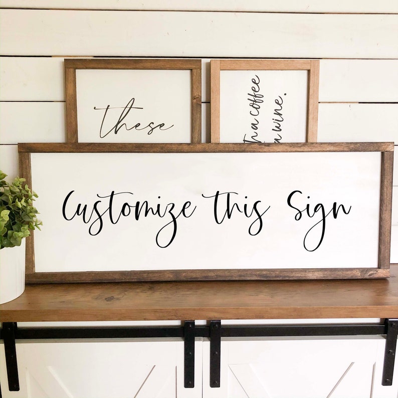 May include: A large rectangular wooden sign with a white background and dark brown frame featuring the black text Customize This Sign. Behind it are two smaller signs, one with the text these and one with the text coffee and wine.