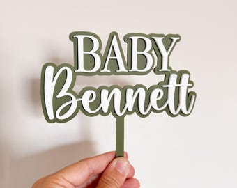 May include: An olive green and white layered cake topper. The top layer reads "BABY" in a serif font, and the bottom layer reads "Bennett" in a cursive font. The topper is held by a thin stem.