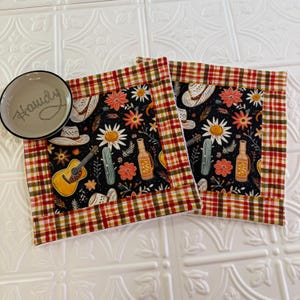 May include: Two square fabric coasters featuring a black background with a pattern of yellow acoustic guitars, white cowboy hats, orange flowers, and green cacti, bordered by a red, orange, and yellow plaid trim, next to a small bowl with "Howdy" text.