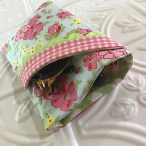 May include: A floral patterned fabric sunglasses case with a pink and white gingham trim and a green scalloped trim. The case is folded closed and has sunglasses inside.