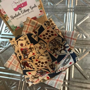 May include: A stack of fabric bowl cozies with a playing card design, plaid, and floral patterns. The cozies are displayed on a textured metal surface. A card with the text "Roadside Cottage Goods" is in the background.