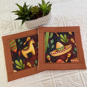May include: Two square fabric coasters featuring a dark background with a print of yellow horses, sombreros, maracas, and cacti. Each coaster has a brown and white gingham border. A small white woven basket contains green succulents.