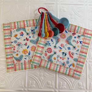 May include: Two square fabric pot holders featuring a white background with a colorful pattern of blue birds, red and yellow flowers, and green leaves, bordered by a pink, green, and white plaid trim, with a set of plastic measuring spoons.