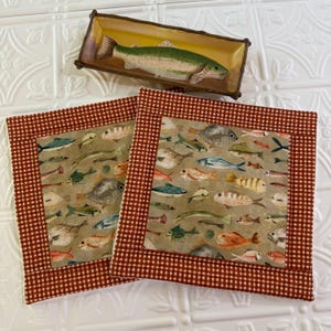 May include: A rectangular ceramic dish featuring a painted green fish, positioned above two square fabric pot holders. The pot holders have a tan center panel printed with various colorful fish and a red and white checkered border.