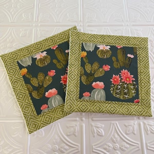 May include: Two square fabric coasters featuring a dark green background with a repeating pattern of green cacti and bright pink flowers. Each coaster has a wide border of light green fabric with a white geometric diamond pattern.