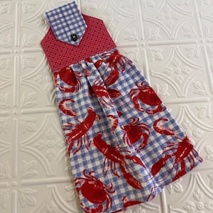 May include: A hanging kitchen towel featuring a blue and white gingham pattern with a red lobster and crab print. The top section is a red fabric with small white stars, a blue and white gingham tab, and a single black button closure.