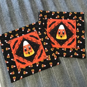 May include: Two black fabric coasters with orange and yellow candy corn designs. The coasters feature a smiling candy corn with a black outline and bats on a black background.