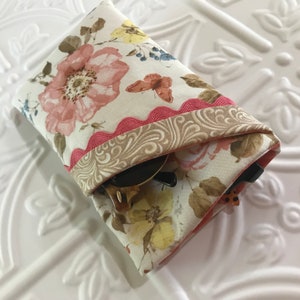 May include: A floral print glasses case with a pink scalloped trim and a decorative beige border. The case features a pattern of pink roses, yellow flowers, and a butterfly. Sunglasses are partially visible inside the case.