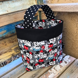 May include: A black and white polka dot fabric tote bag with a black fabric handle. The tote bag is lined with black fabric and features a gray fabric exterior with a repeating pattern of Mickey Mouse and Minnie Mouse.