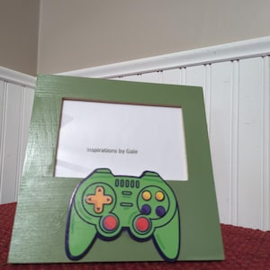 May include: Green picture frame with a painted video game controller design. The controller is green with orange, red, and purple buttons. The frame has the text "Inspirations by Gale".