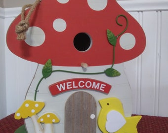 May include: A wooden birdhouse shaped like a red and white mushroom with a small door and the word "WELCOME" on the front. The birdhouse has a yellow bird with a white heart on its chest and two yellow mushrooms with white dots on the side.