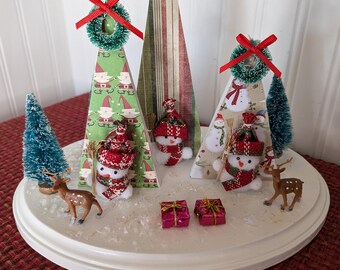 May include: A Christmas-themed centerpiece featuring three paper cone trees adorned with wreaths and red bows. The base includes snowmen, miniature deer, small wrapped gifts, and bottle brush trees, all on a white, round base.