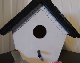 May include: A white birdhouse with a black roof and a decorative trim. The birdhouse has a small bird figurine and a pink flower pot on the base.