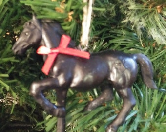 May include: A black horse ornament with a red bow, hanging from a natural twine string. The horse is posed mid-stride, with detailed features. The ornament is set against a backdrop of green pine needles.