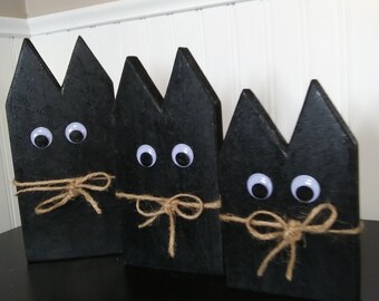 May include: Three black wooden cat figures with googly eyes and twine bows. The cat figures are arranged in a row, each with a different height. The background is a white wall.