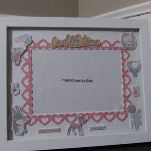 May include: White picture frame with a pink and white heart border. The frame has a cutout for a photo and includes various cutouts of animals and the words "cuddle time", "sweet baby", and "cuddle repeat".